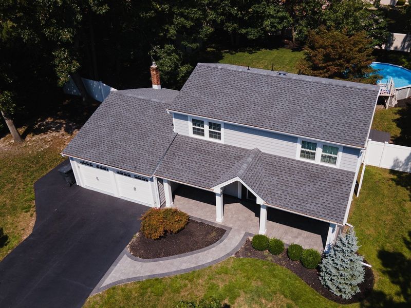 Aerial view of a gray-roofed house with a black driveway and a pool in the background.