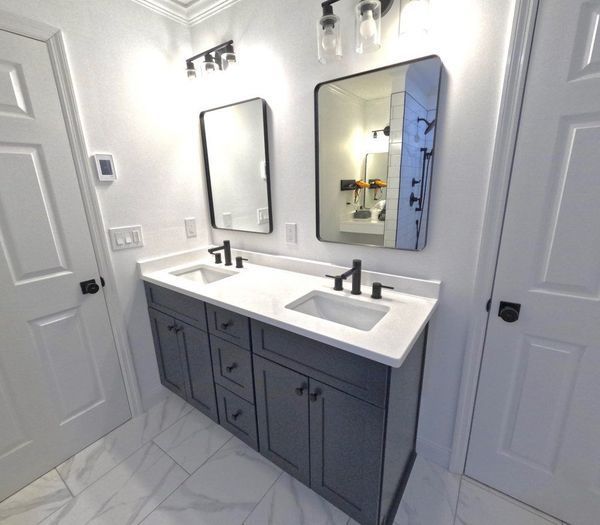 Bathroom with double vanity, two mirrors, and gray cabinets. White countertop, black fixtures, and white tile flooring.