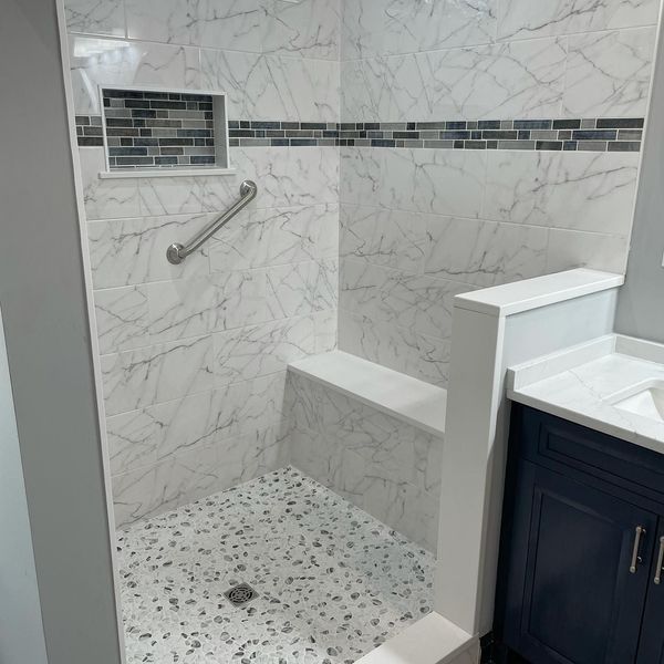 White marble-tiled shower with a bench, handrail, and mosaic accent. A built-in niche and a pebble-stone floor.