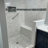 Accessible shower with built-in seat, grab bar, and stone floor. White marble-like tile with a dark mosaic accent.