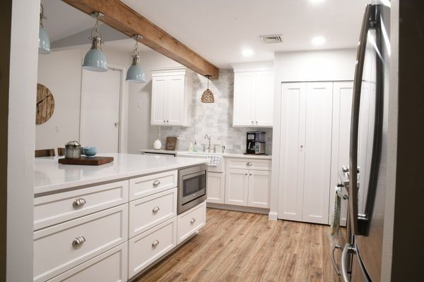 White kitchen with island, cabinets, and appliances. Wooden floor and ceiling beam, pendant lights.