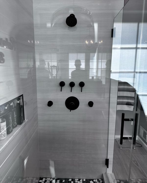Modern shower with black fixtures on gray-tiled wall, glass door, and a built-in shelf.