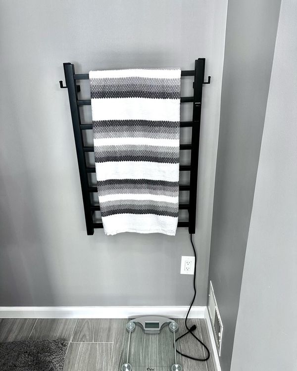 Black towel warmer with a striped towel on a gray wall, near a scale and power outlet.