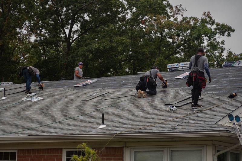 Roofers working on a gray shingled roof, trees in the background, cloudy sky.