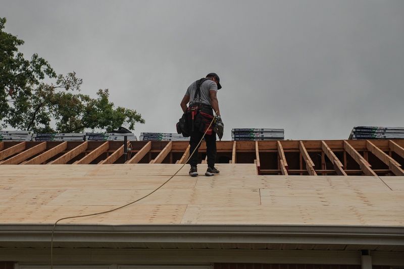 Roofer working on a wooden roof framework on an overcast day; safety harness attached.