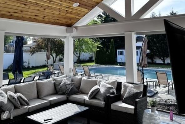 Covered patio with sectional sofa, overlooking a pool and yard. White beams, wood ceiling, outdoor TV.