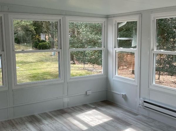 Sunroom interior with multiple windows, white walls, and wood-look flooring overlooking a backyard.