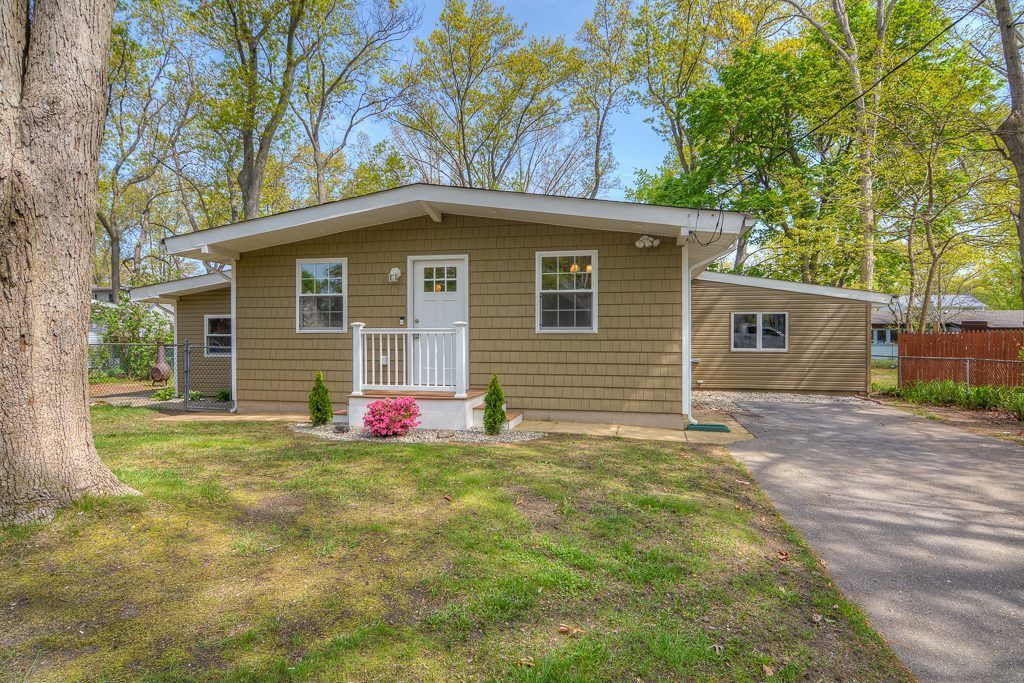 Small beige bungalow house with a white front door and two windows. Green grass and trees surround it.