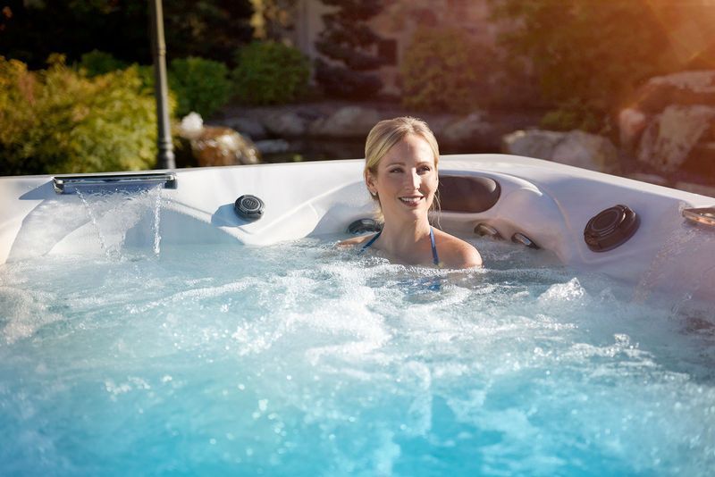 Blonde woman smiling in a bubbling outdoor hot tub on a sunny day.