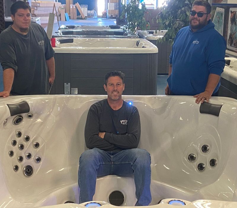 Three men in a hot tub showroom. One sits in the tub smiling, while two others stand behind.