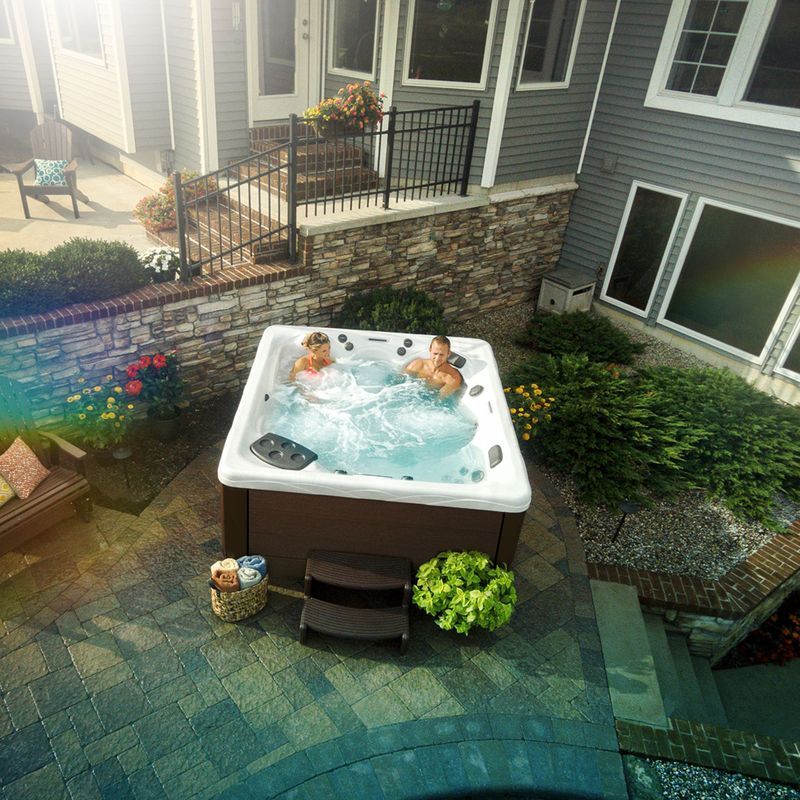 Two people relax in a hot tub on a patio near a house with sunlight.
