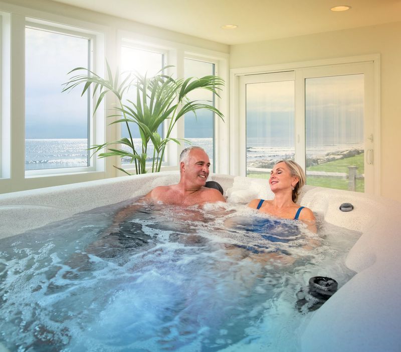 Mature couple relaxing in a hot tub with a coastal view. Smiling, sunny interior.