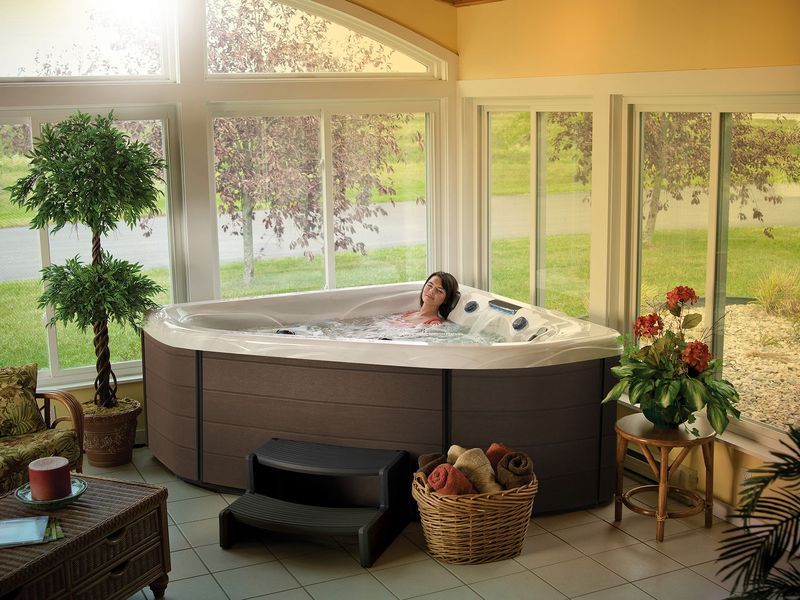 Woman relaxing in a triangular hot tub in a sunroom with a view of trees.