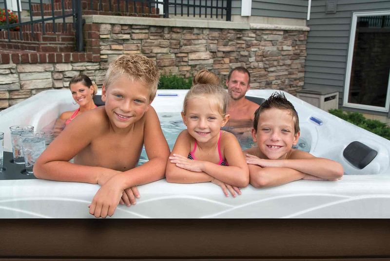 Family smiling in a hot tub; two boys, girl, and parents, brick and siding background.