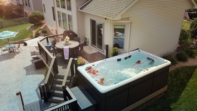 People enjoying a swim spa on a deck, next to a house with a yard and outdoor furniture.