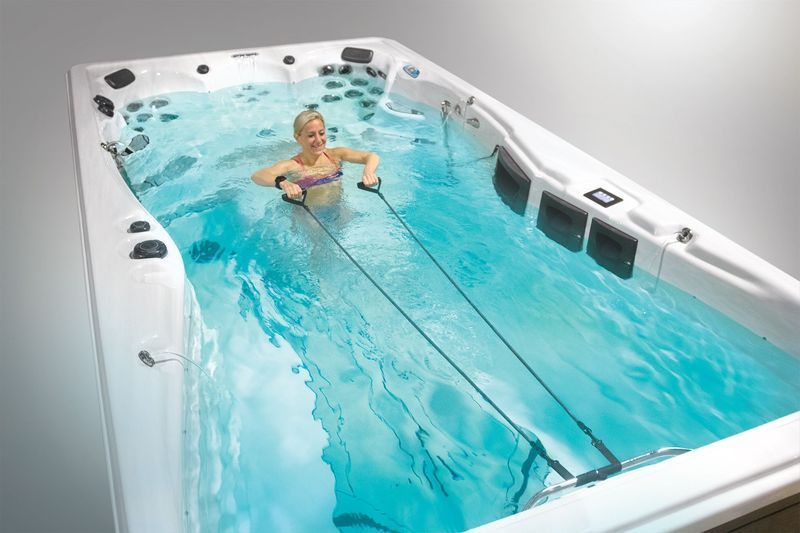 Woman exercising in an aquatic fitness pool, pulling resistance bands. Light blue water, white pool, neutral background.