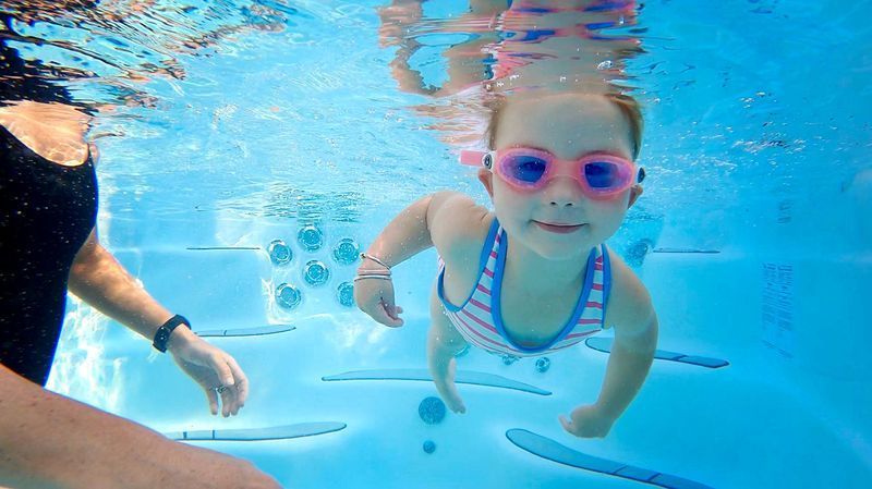 Girl in goggles swims underwater in a pool, smiling towards the camera.