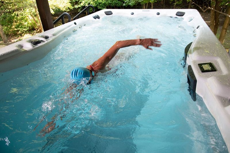 Person swimming freestyle in a swim spa, blue water, sunny outdoor setting.