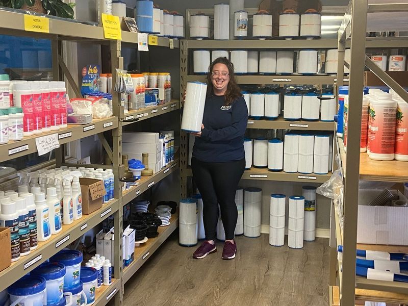 Woman smiles, holding a large filter in a store aisle. Shelves stocked with pool supplies.