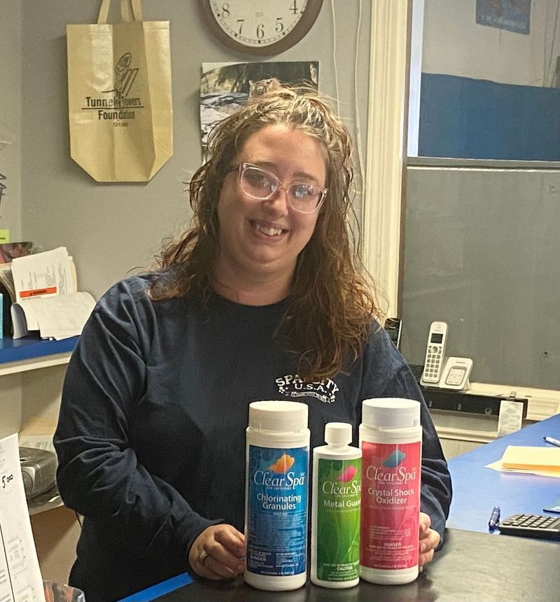 Woman in blue shirt smiles, holding pool chemicals behind a counter.