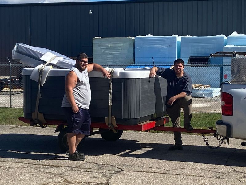 Two men by a trailer carrying two hot tubs; near a warehouse.