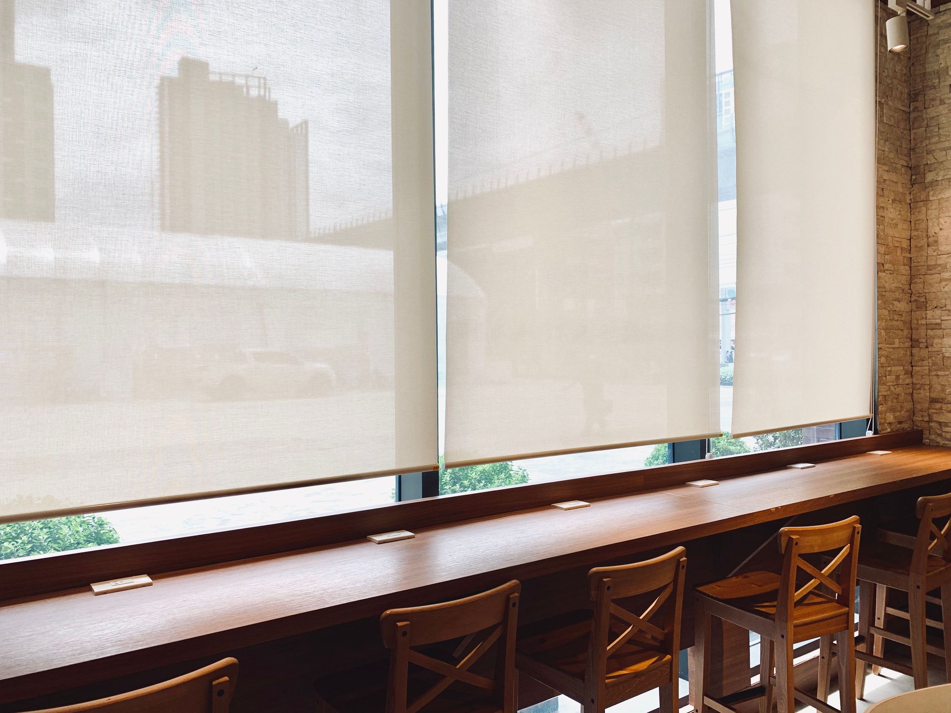 A row of wooden chairs sits at a long counter against a large window covered by light, semi-transparent roller shades.