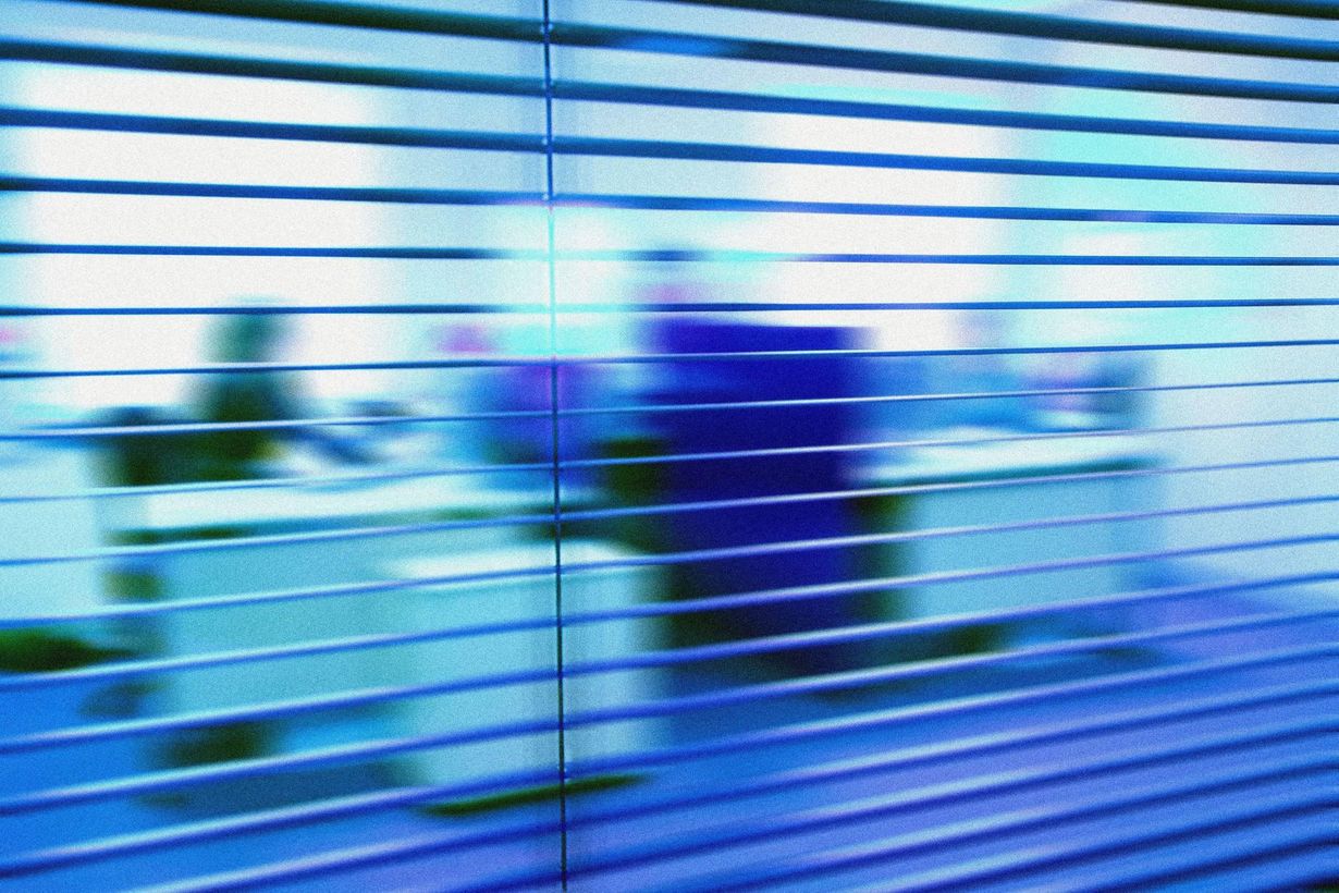 Blurred view through horizontal window blinds of a modern office space with desks and chairs.