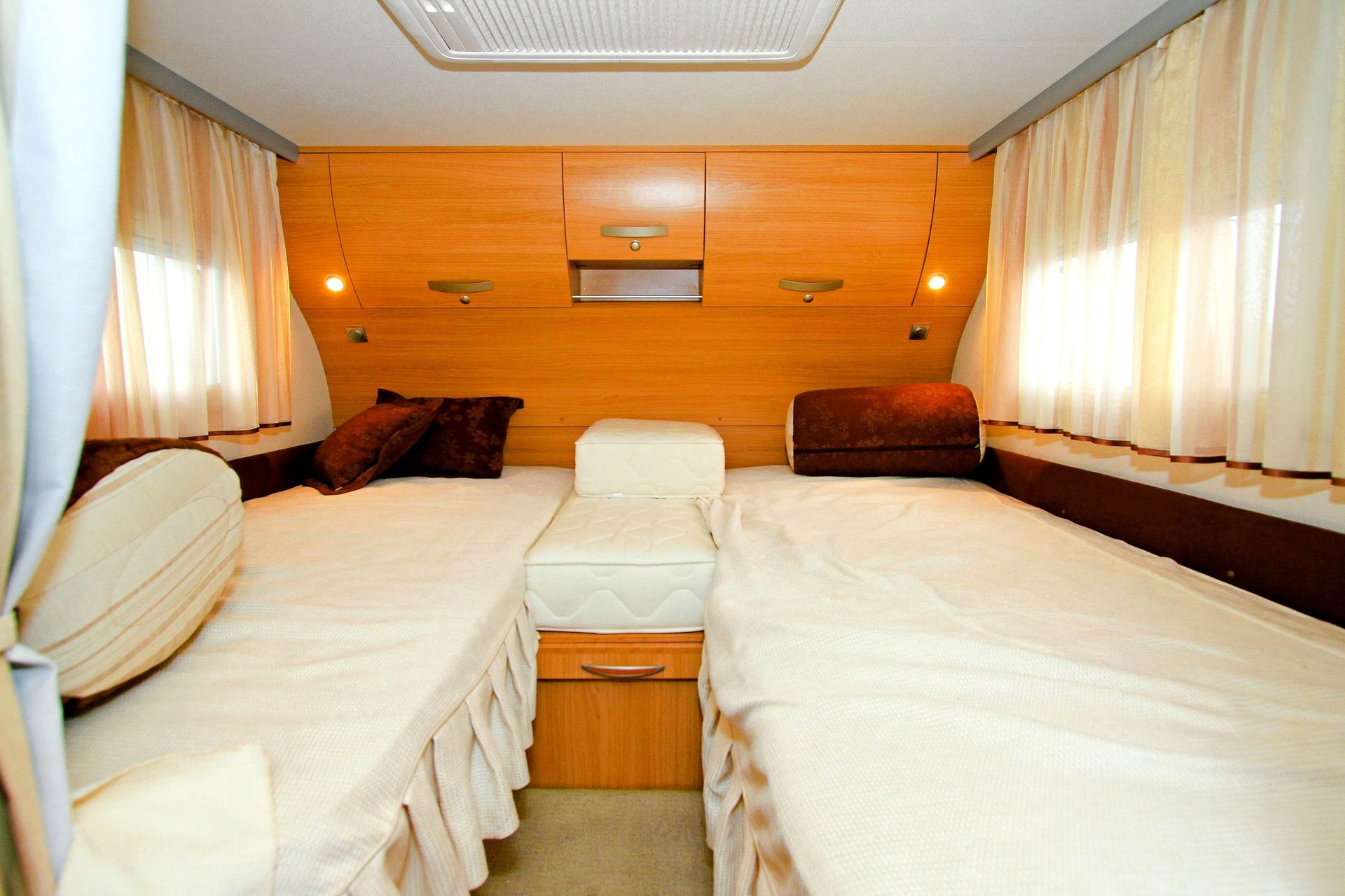 A bright RV interior features two twin beds with white linens, brown pillows, and wood cabinets against the back wall.