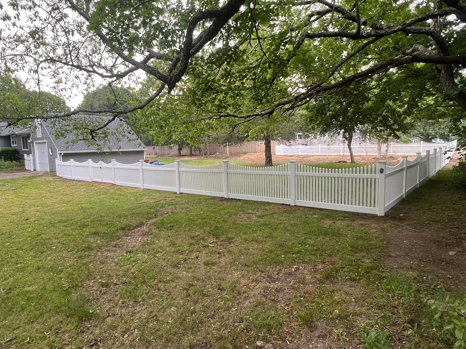 White picket fence encloses a grassy yard, under a large tree. A building is visible behind the fence.
