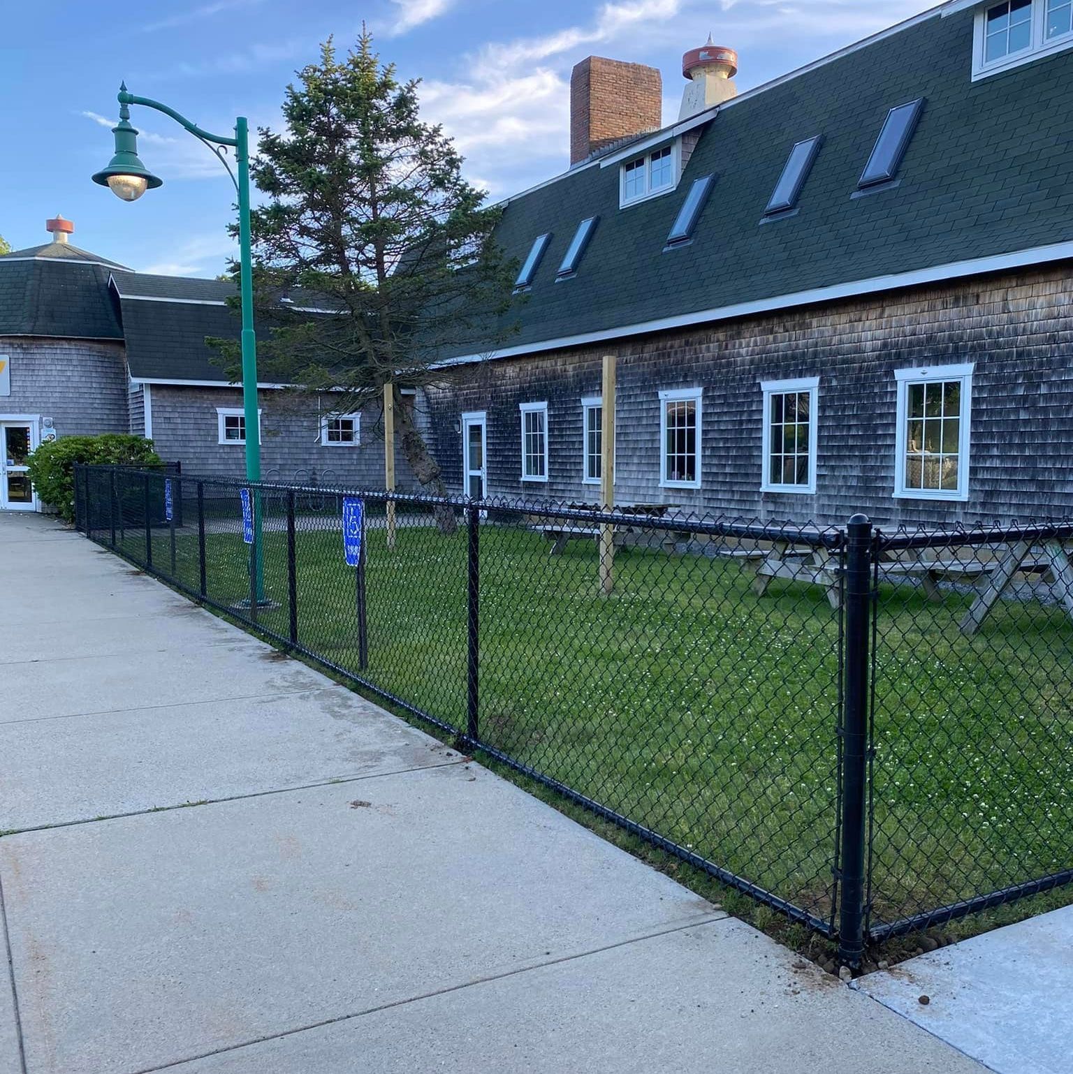 Exterior view of a weathered wooden building with a green roof, windows, and a black chain-link fence bordering a grassy area.