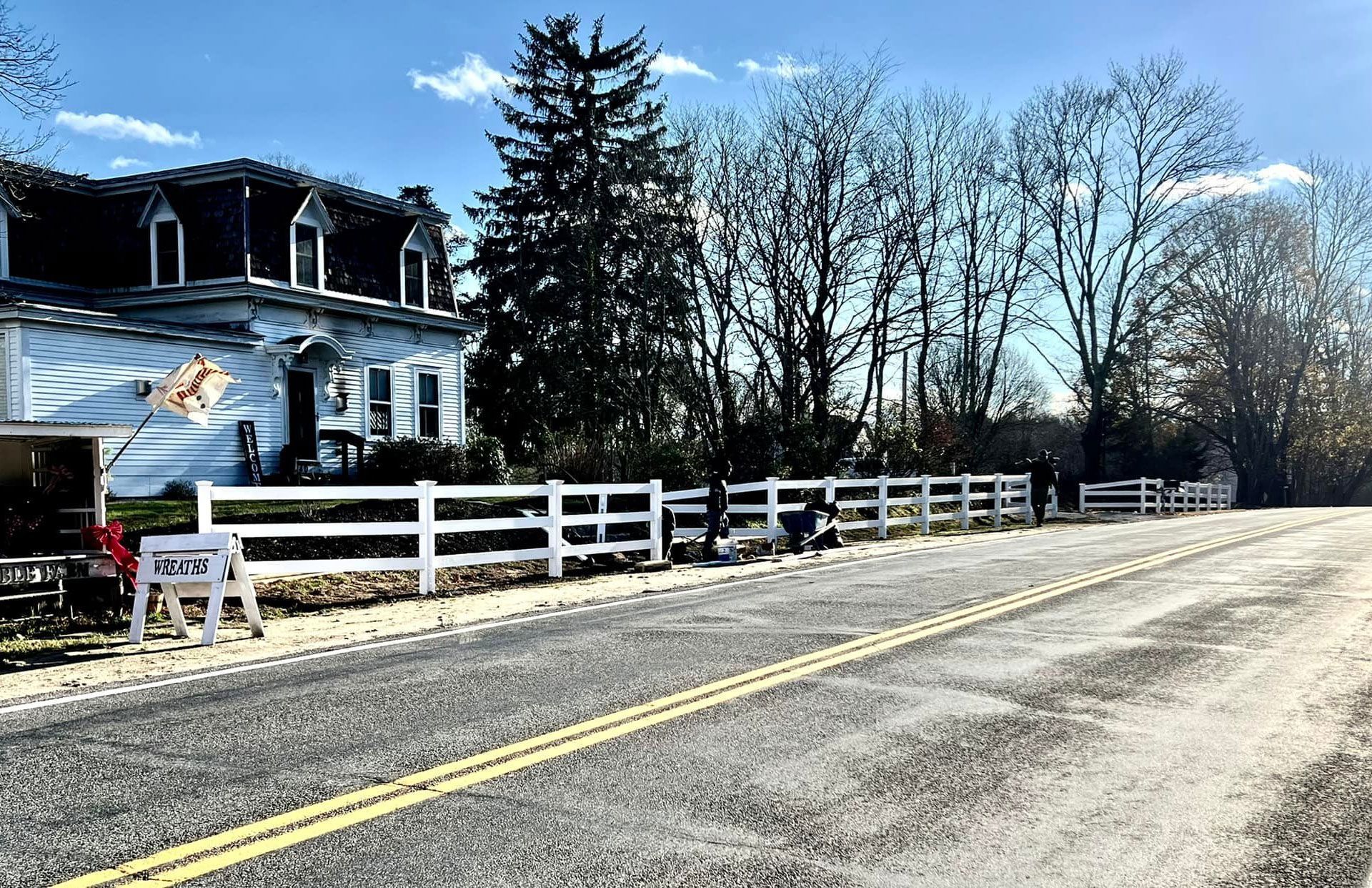 White fence lines a road with a two-story building on the left. Bare trees and a cloudy sky.