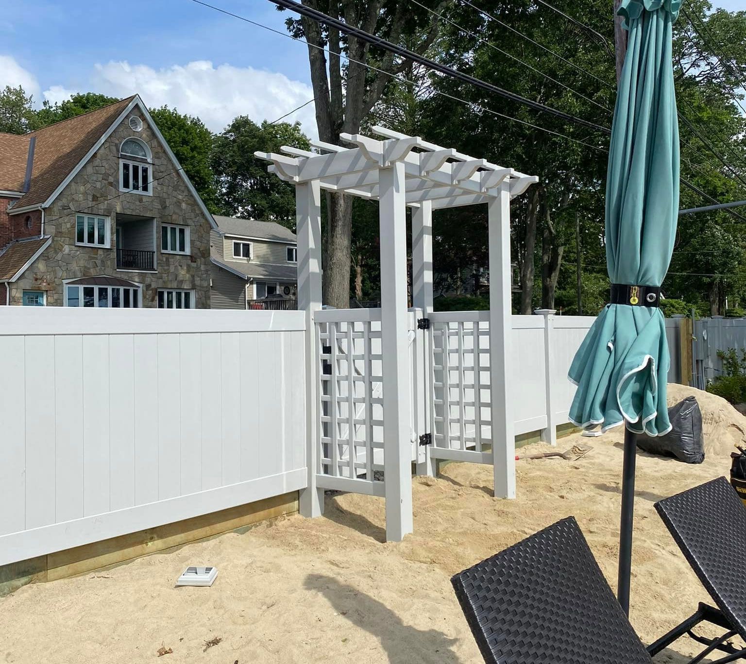 White pergola gate in a white fenced yard, next to a beach umbrella and lounge chairs.