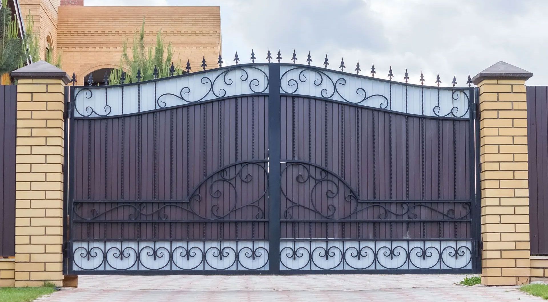 Brown metal driveway gate with decorative ironwork, set between brick pillars, cloudy sky.