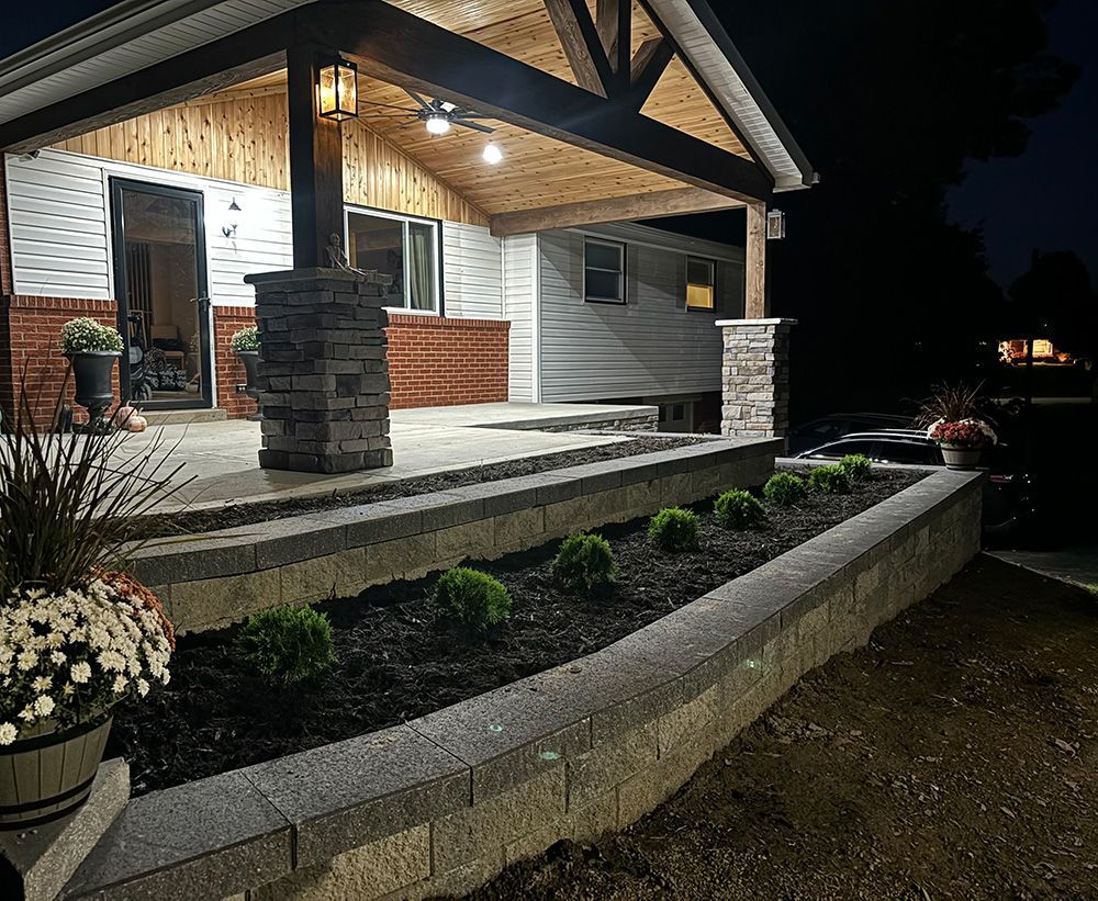 A house porch at night featuring stone retaining walls, dark mulch, small shrubs, and potted flowers under warm lighting.
