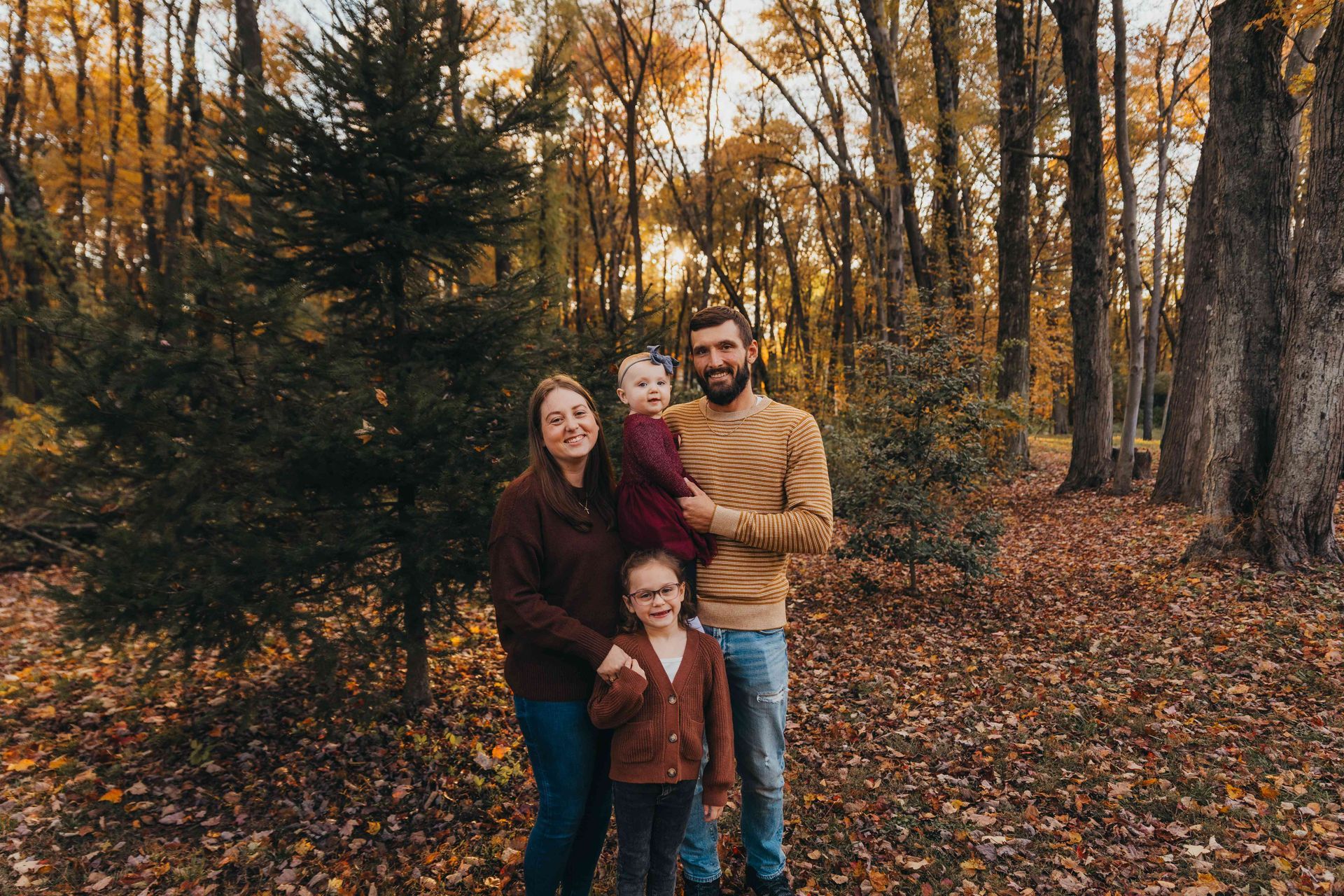 A family of four poses in a wooded area during autumn, surrounded by fall foliage and fallen leaves.