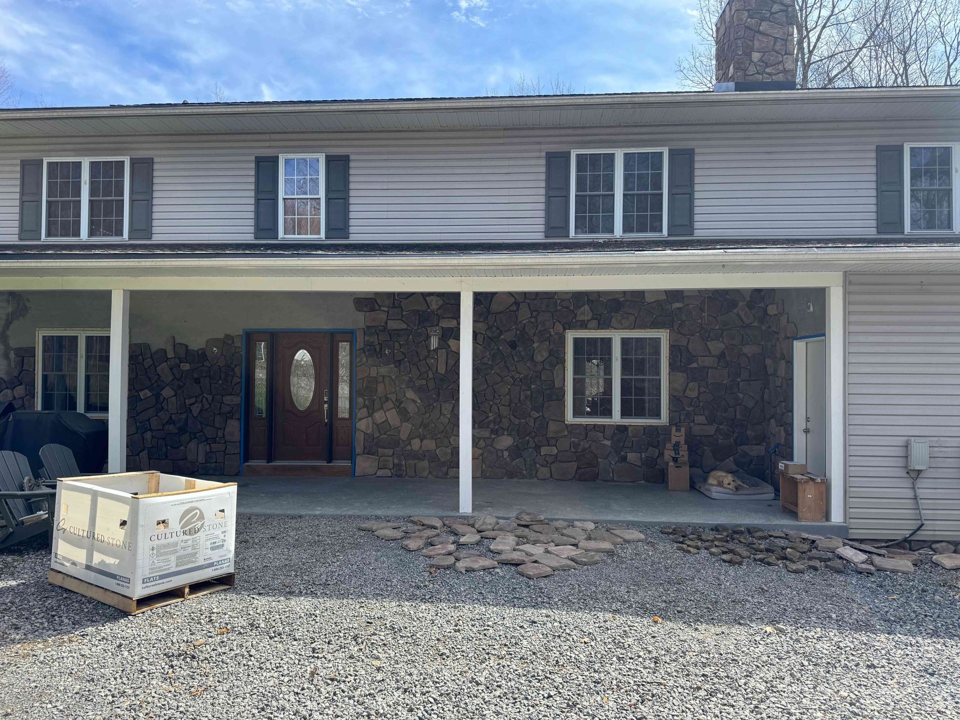 A light-gray two-story house with a rock-faced front porch, a dark wooden door, and a white shipping crate on gravel.