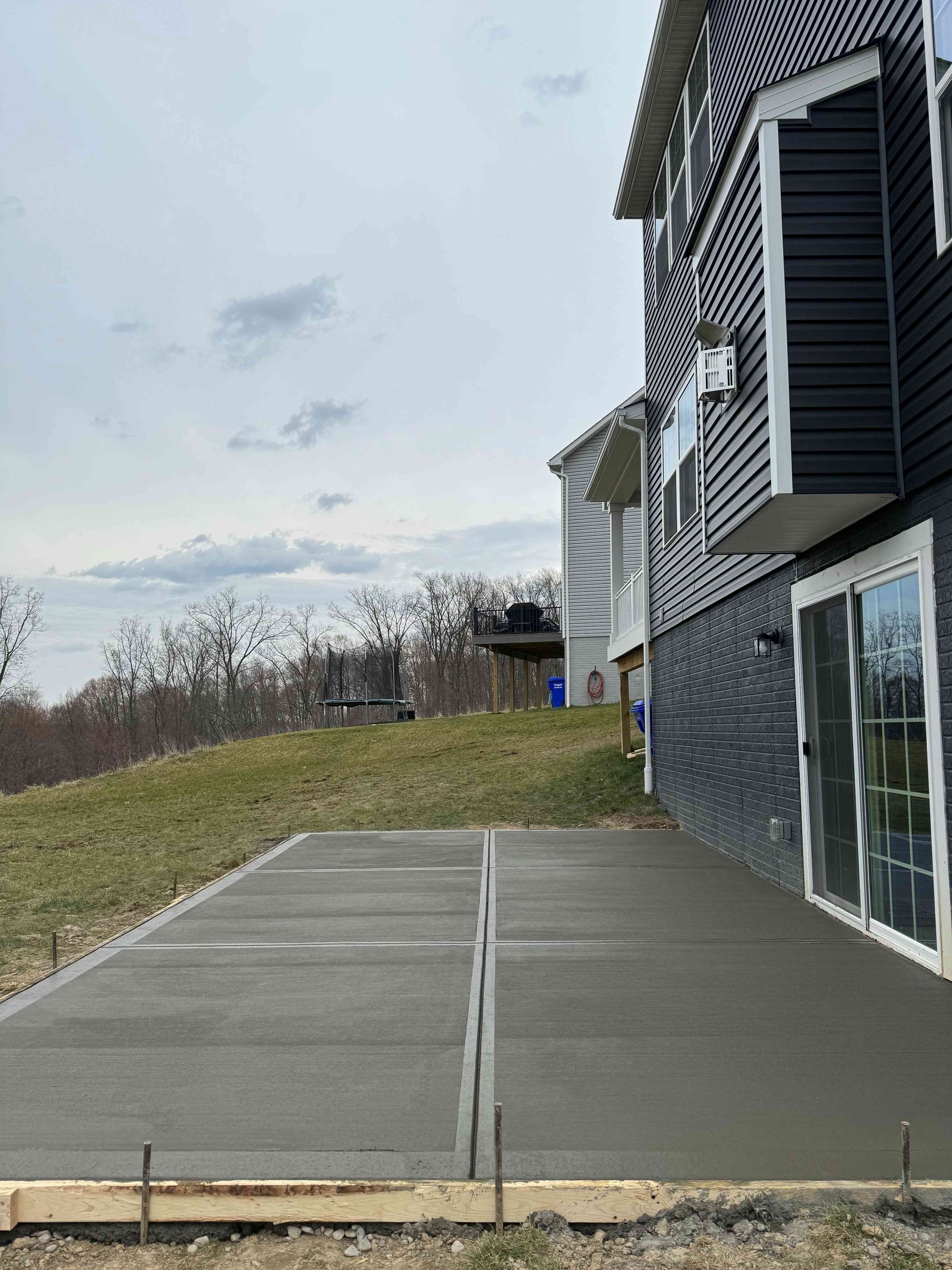 A freshly poured concrete patio adjacent to a gray-sided house on a grassy, sloped hill under a cloudy sky.