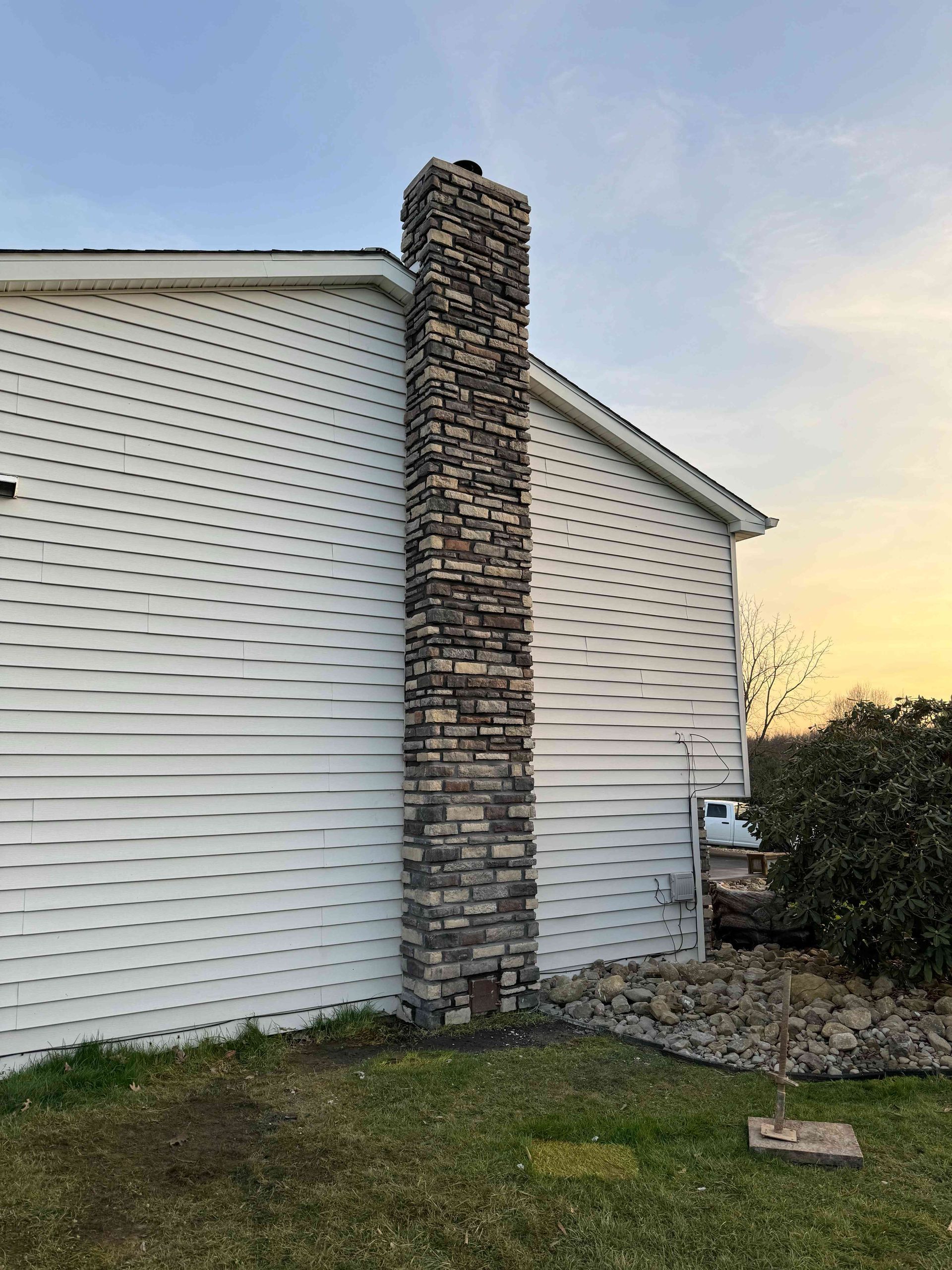 A stone chimney rises against the white vinyl-sided exterior wall of a house at sunset, with grass and gravel below.