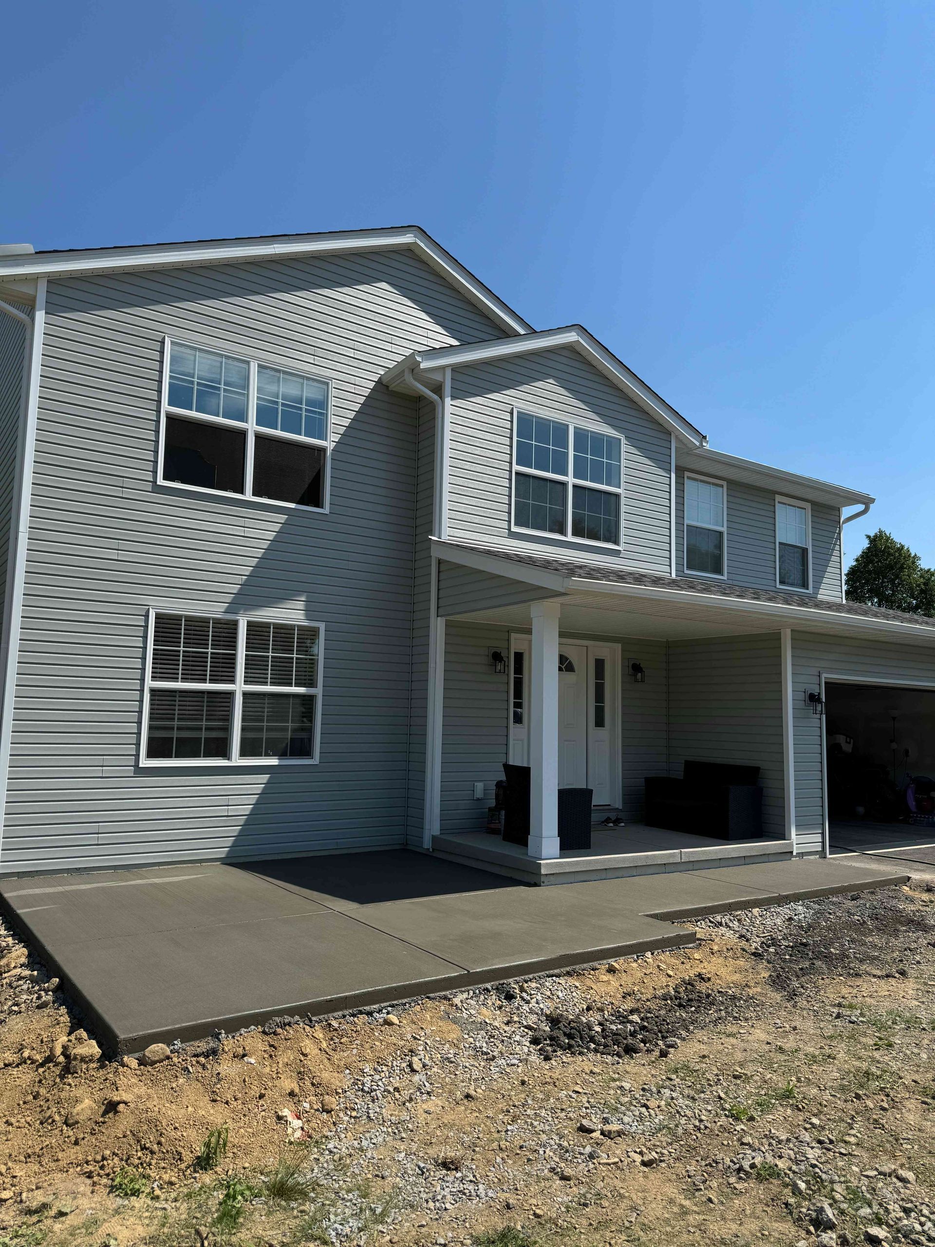 A two-story light gray house with new concrete patio, siding, and a covered porch under a clear blue sky.
