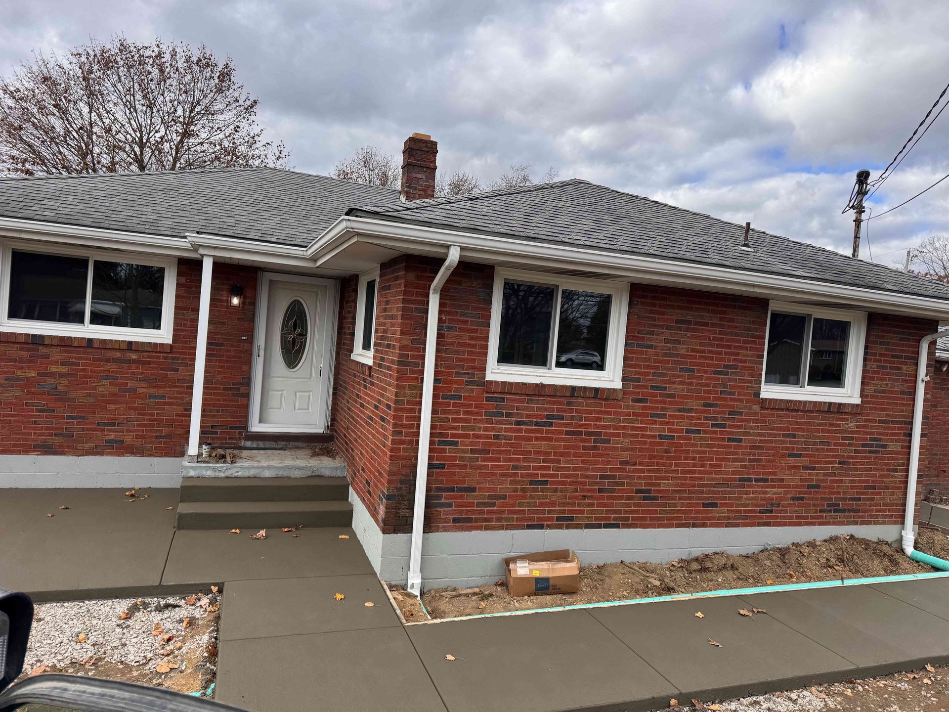 A one-story brick house with a gray shingled roof, a white front door, and a freshly poured concrete walkway.