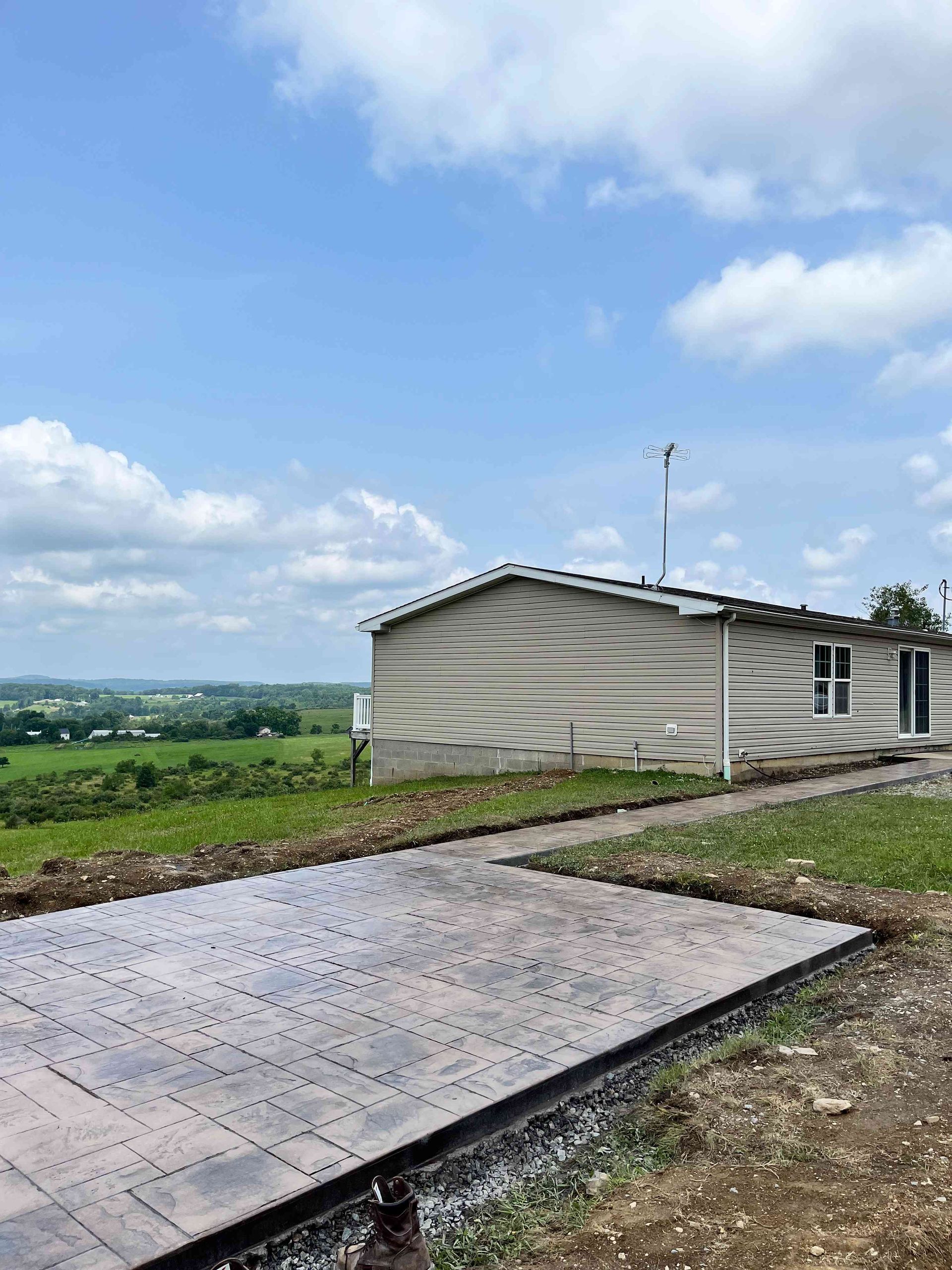 A new light-colored patio paver installation next to a beige house on a grassy hill under a bright, cloudy blue sky.