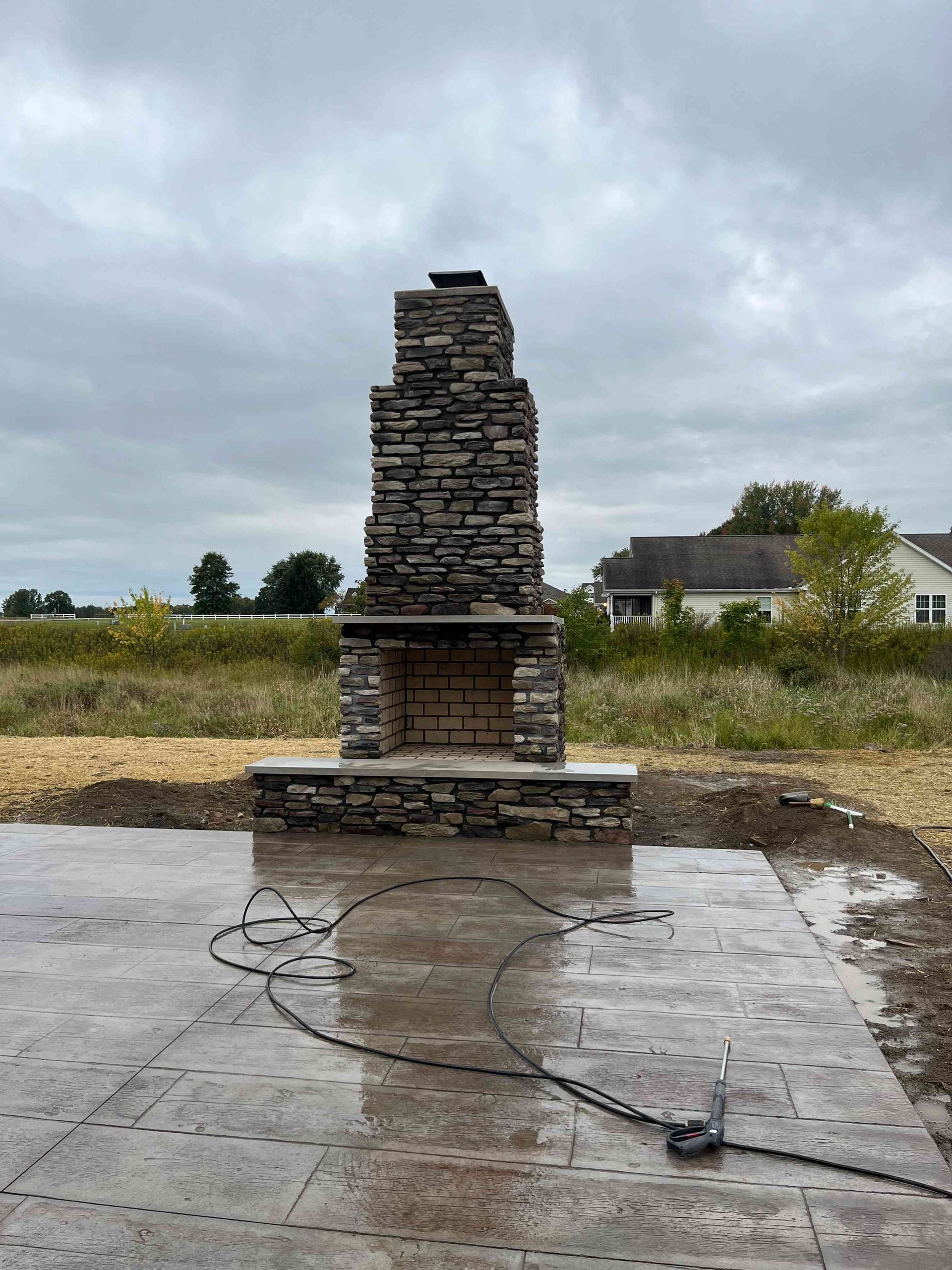 A stone outdoor fireplace stands on a wet concrete patio in a yard against a cloudy sky.