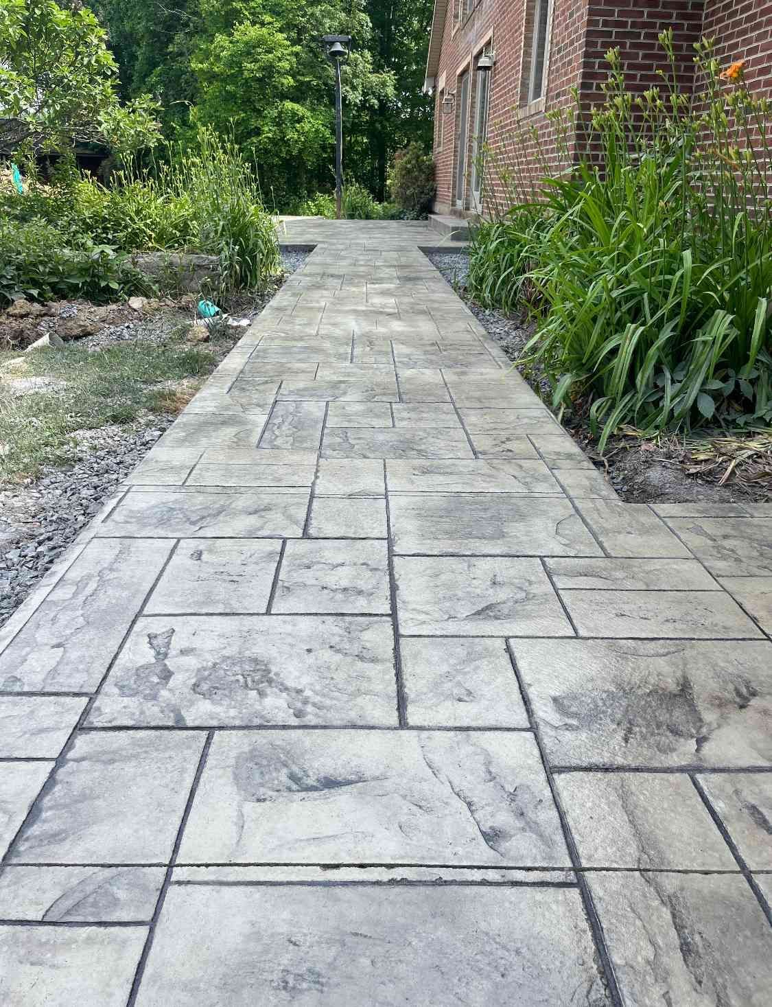 A gray, rectangular-patterned stone walkway leads toward a brick building, surrounded by greenery.