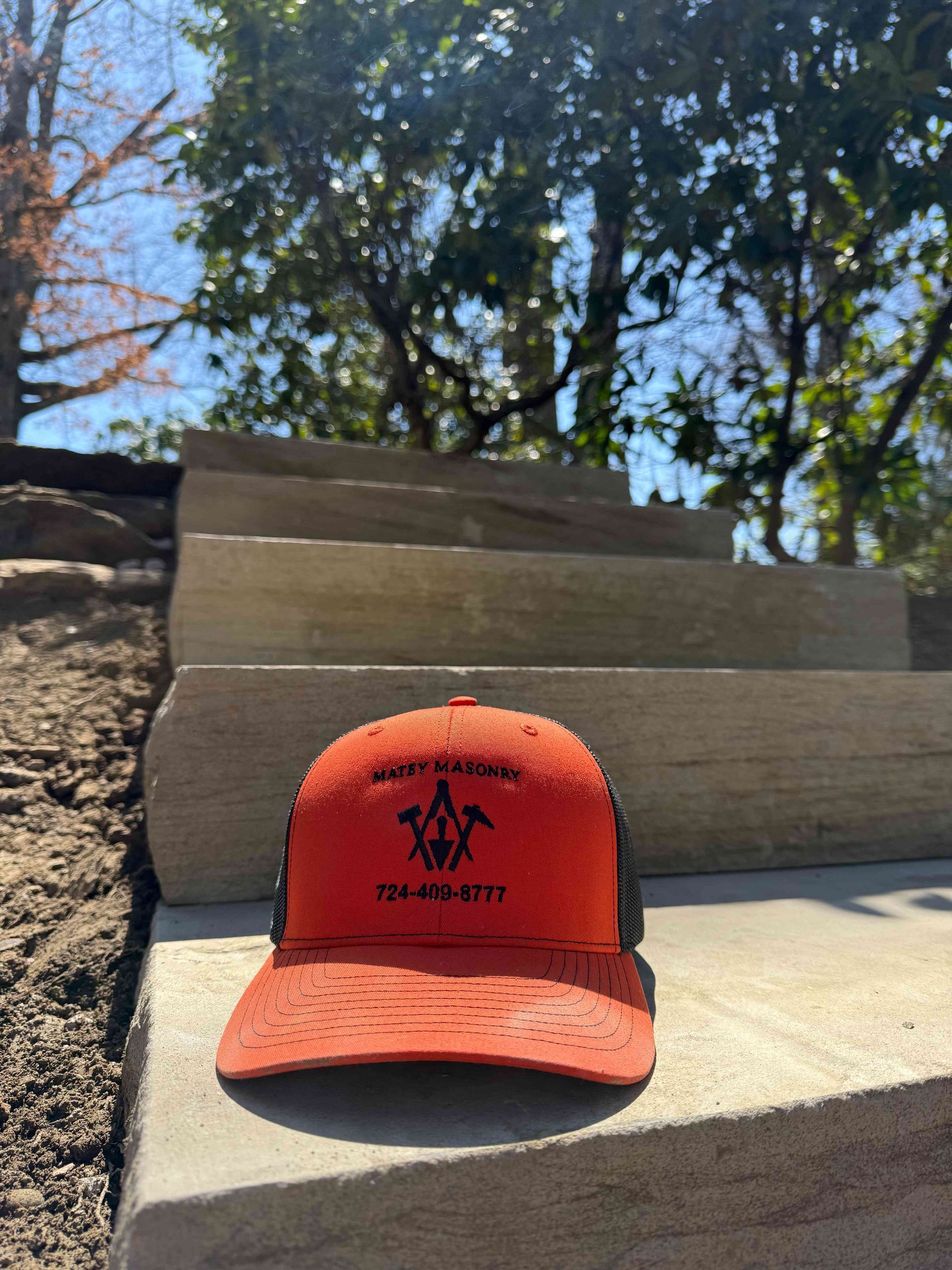 An orange, distressed-style trucker hat sits on a stone step outdoors, featuring a black logo and phone number.