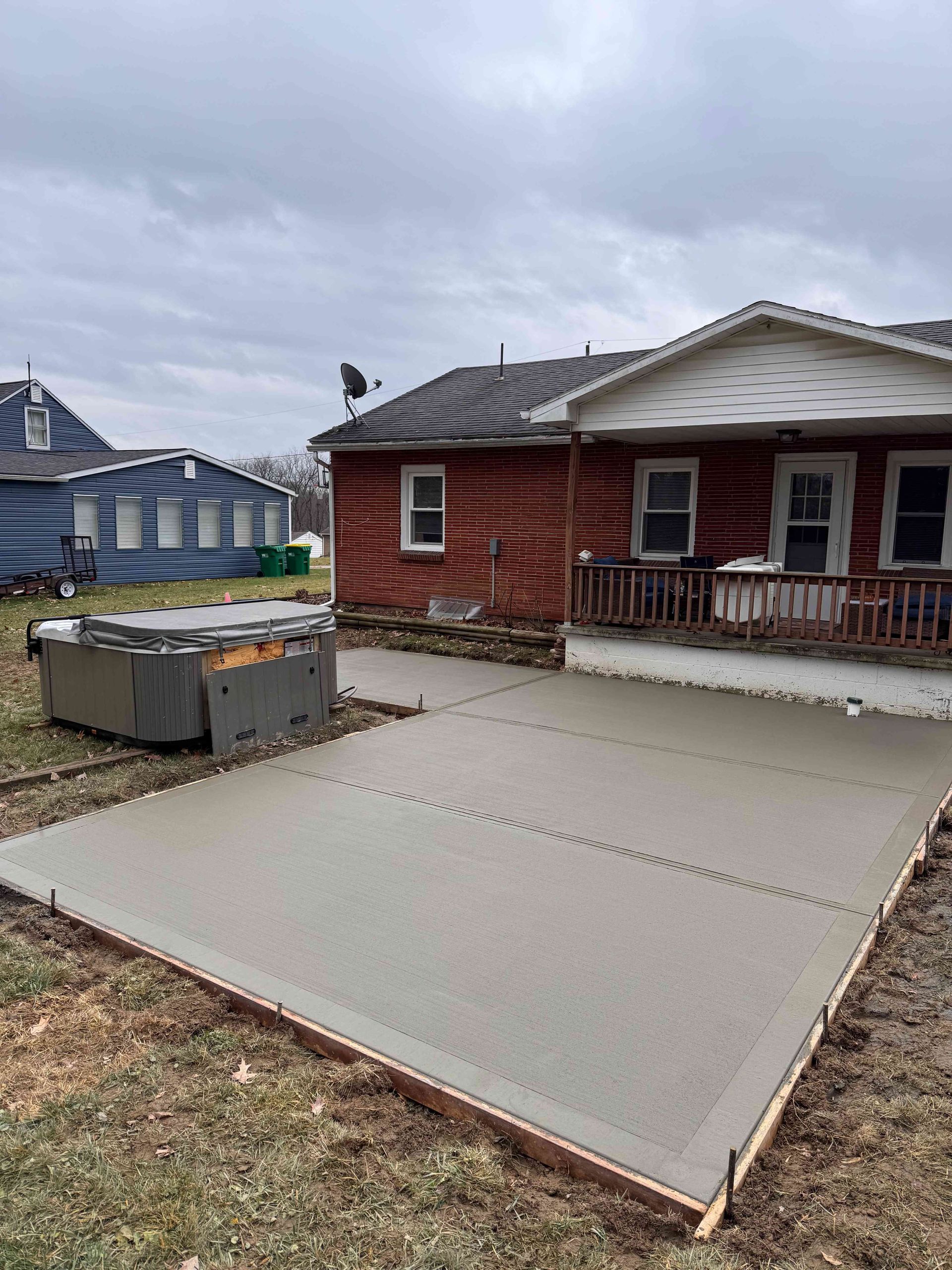 A newly poured concrete patio in a yard next to a red brick house under a cloudy sky.