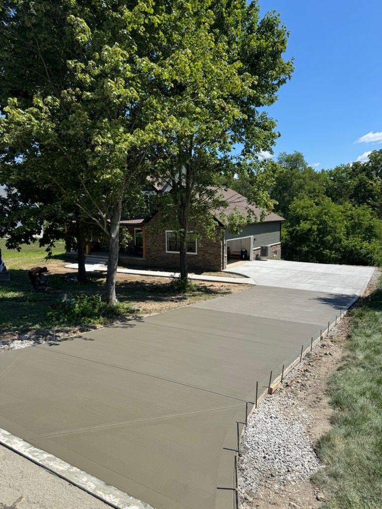 Fresh concrete driveway leading to a house with a garage, bordered by trees and gravel under a blue sky