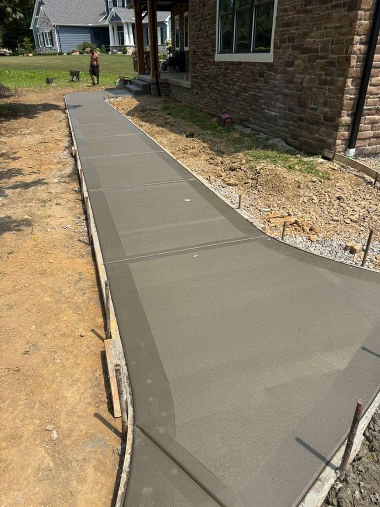 Fresh concrete sidewalk beside a brick house under construction, with dirt along the edges