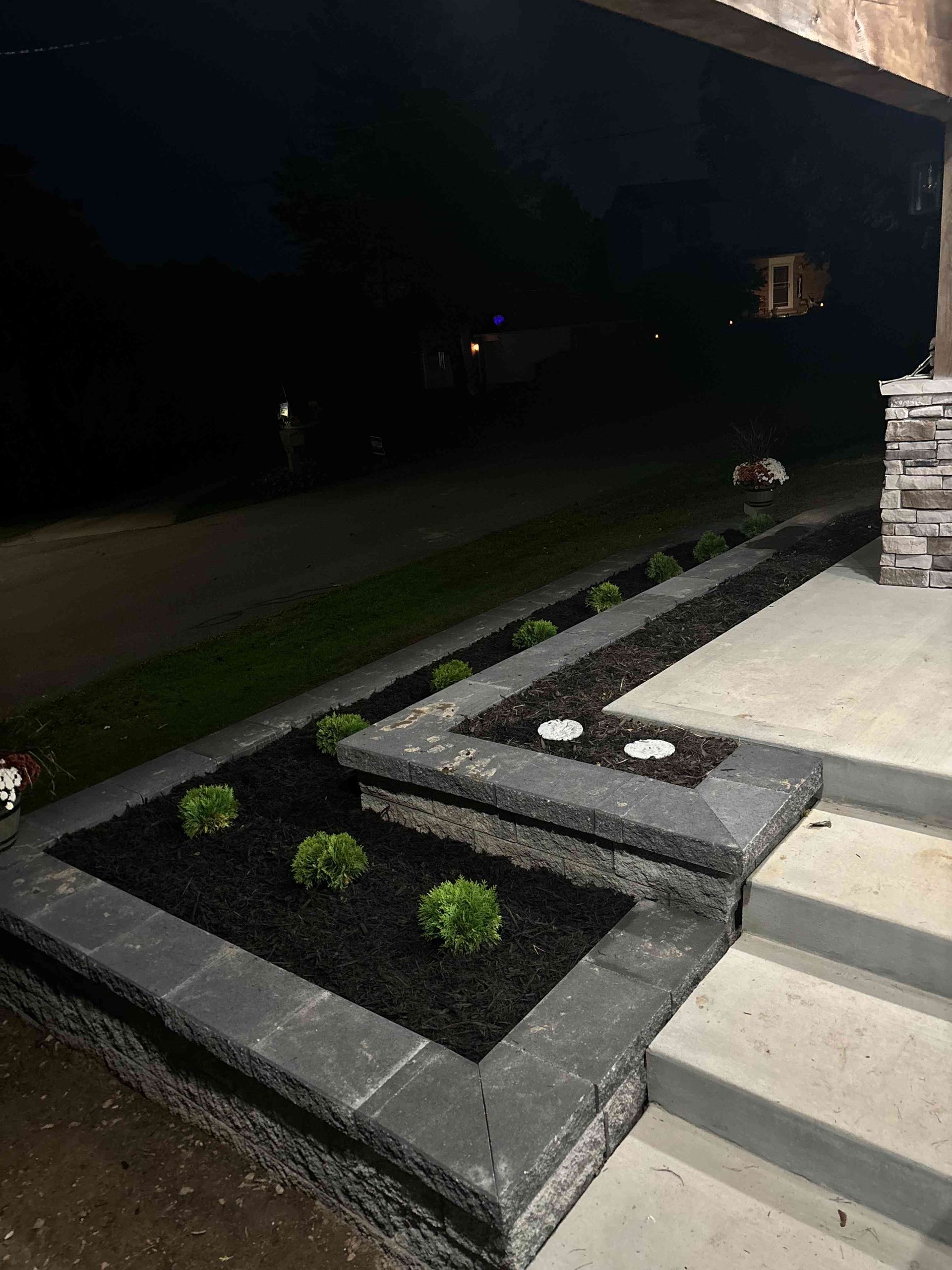A tiered stone retaining wall with black mulch and small shrubs bordering a concrete porch at night.
