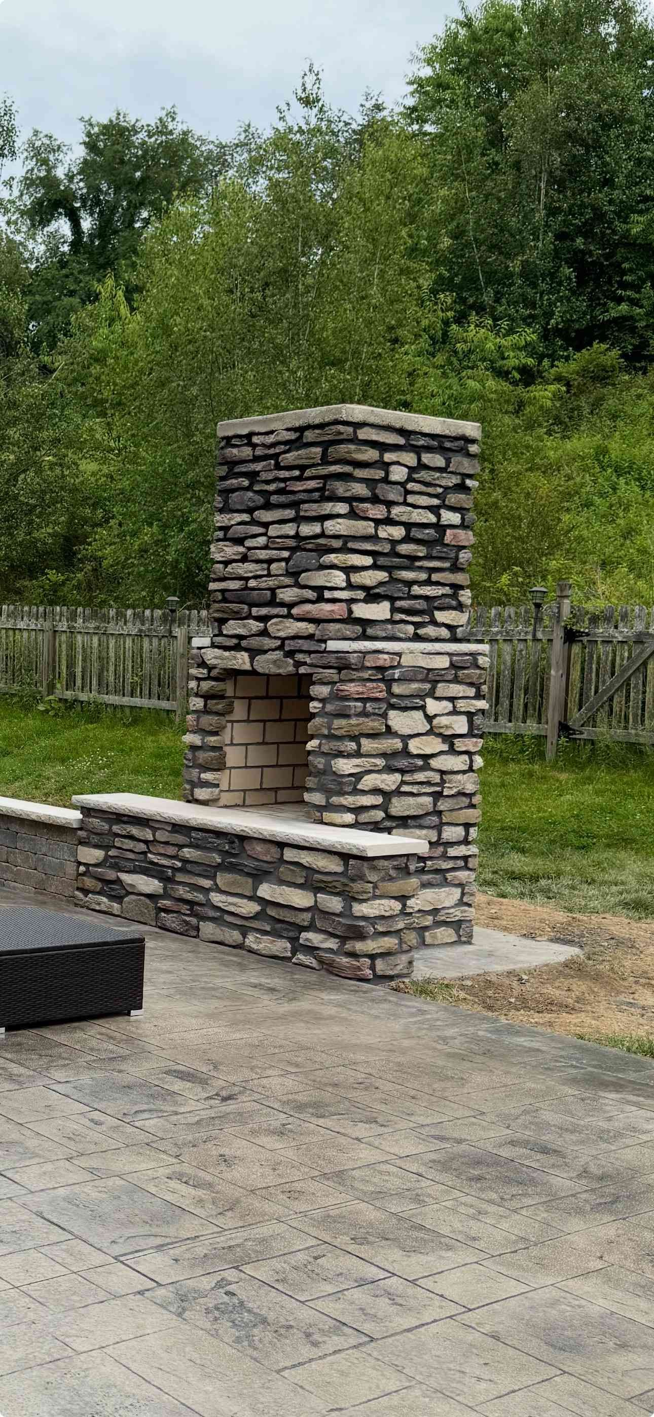 A rustic stone outdoor fireplace stands on a patio, featuring a light brick firebox and stone mantel against a garden.