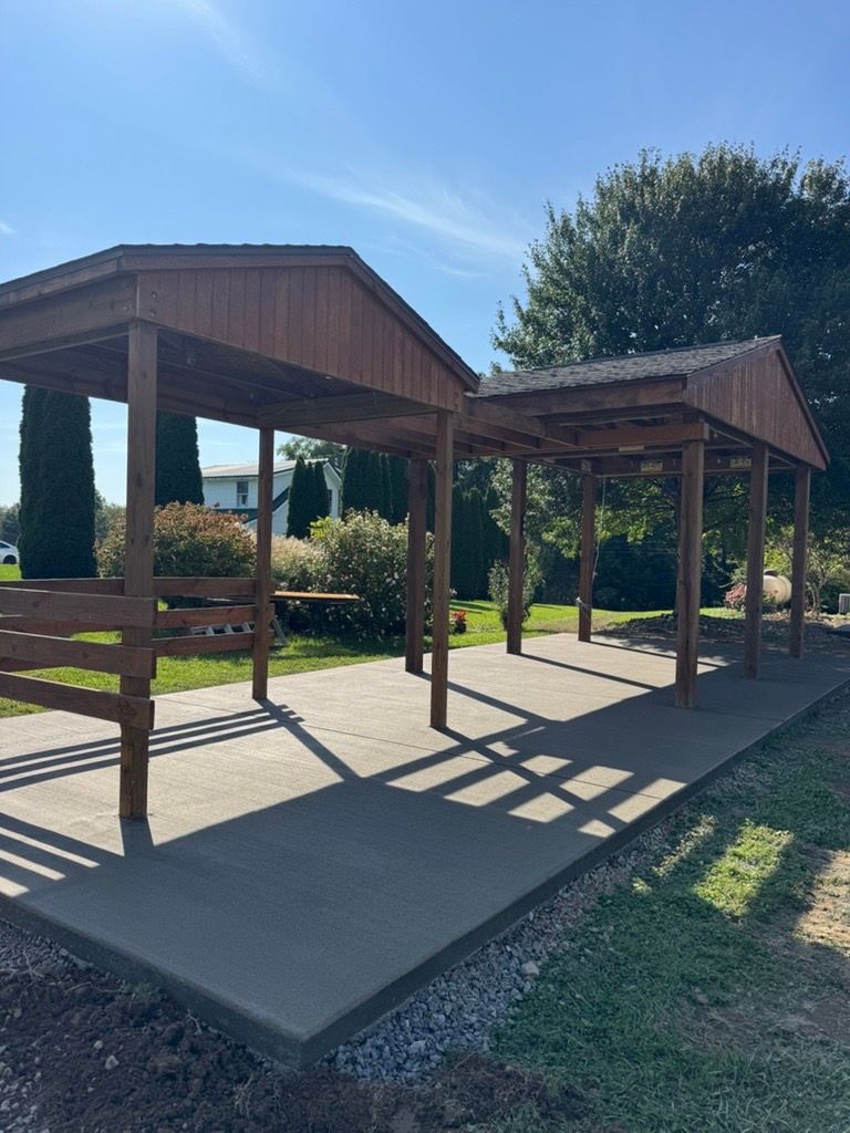 Wooden picnic shelter with benches in a sunny park, casting long shadows on a concrete pad.